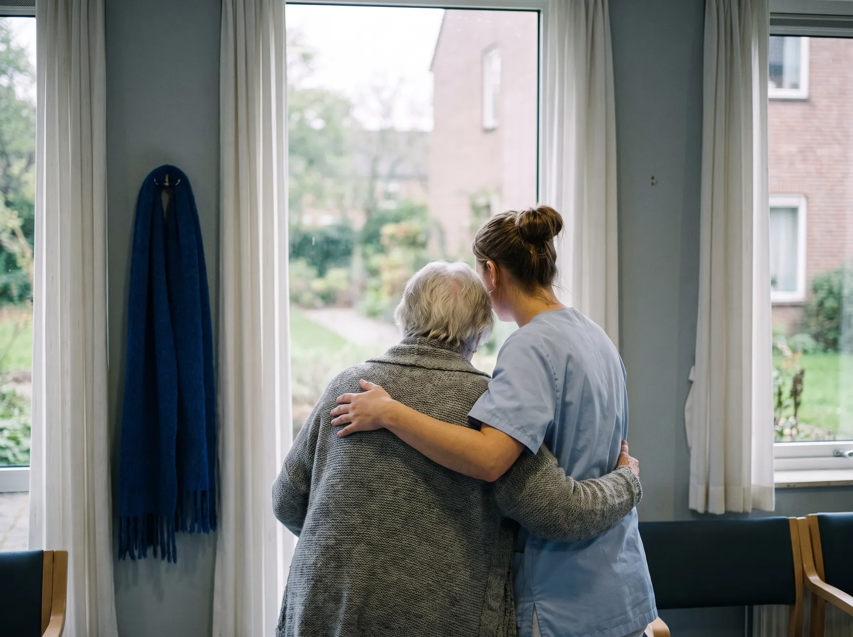 A carer standing with her arm around a resident at the window of a care home
